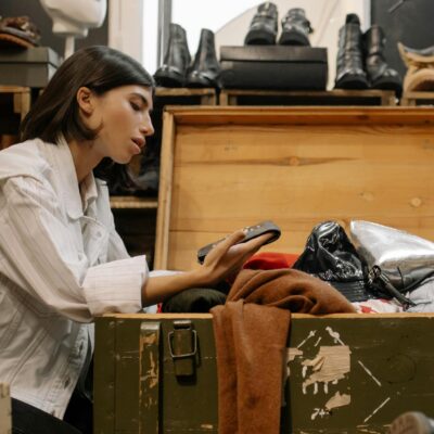 A woman examines clothes from an open chest in a thrift store filled with shoes.