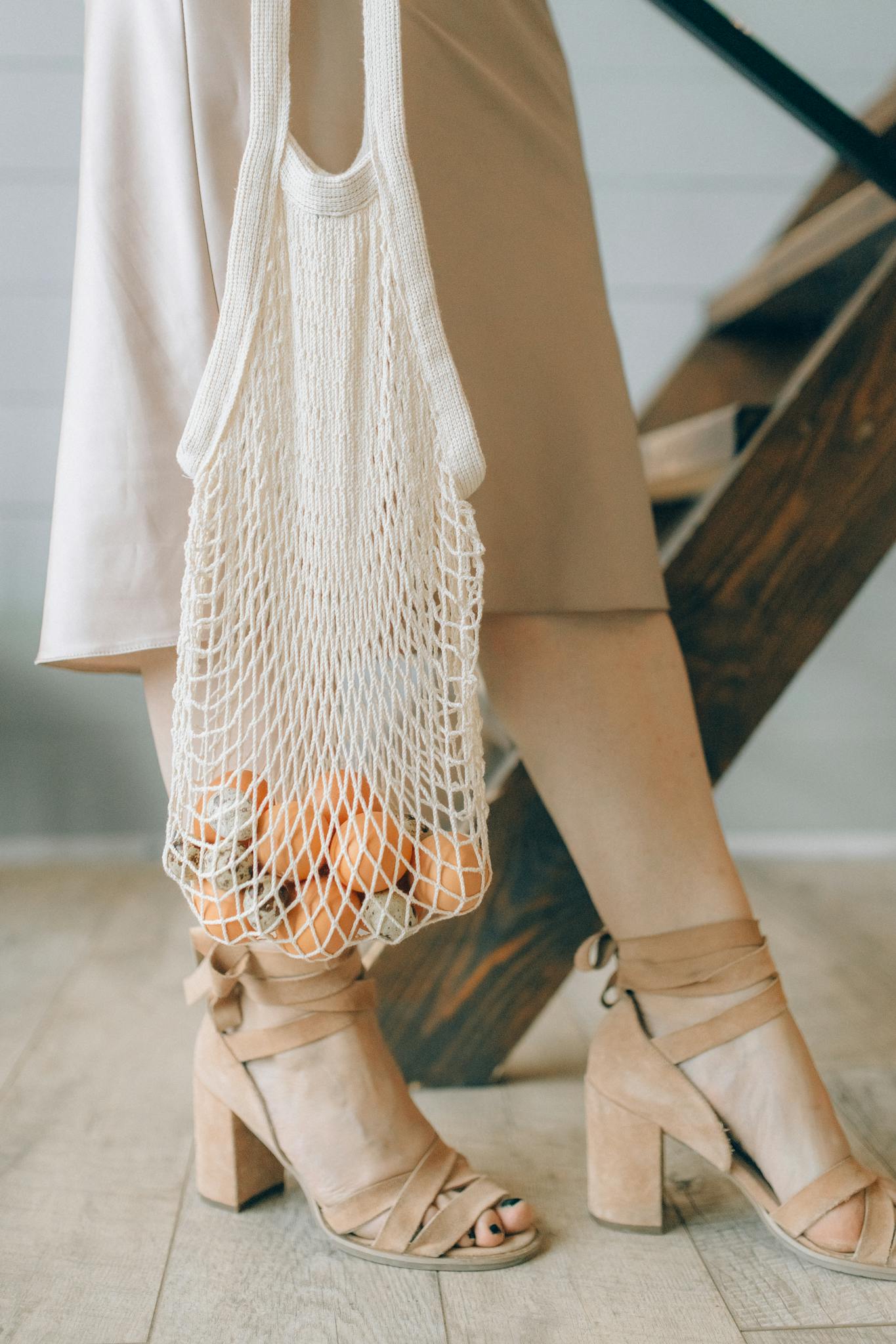 Close-up of a woman in heels carrying a net bag filled with groceries, promoting eco-friendly fashion.